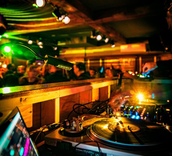 Close-up of a DJ's setup in a dimly lit club with colorful lights, as the crowd vibrates to the beat. A turntable, mixer, and laptop are in focus, with the DJ's hand deftly adjusting controls. The backdrop reveals people enjoying cocktails at the bar and grill, adding to the lively atmosphere.