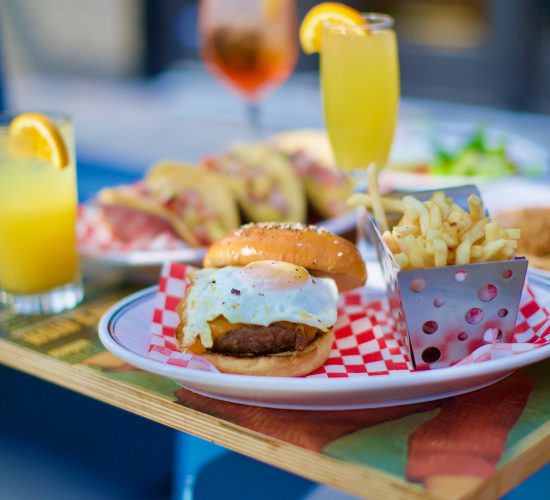 A table showcases a meal featuring the best burger topped with a fried egg, a basket of fries, and a yellow drink with an orange slice. Additional drinks and food items are in the background, including a taco and another drink garnished with an orange slice.