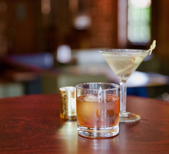 A close-up of a round wooden table holding three items: a glass with an amber-colored drink and ice, a martini glass with a clear cocktail garnished with an olive, and a small golden candleholder. The background of this best restaurant scene is an out-of-focus and dimly lit room.