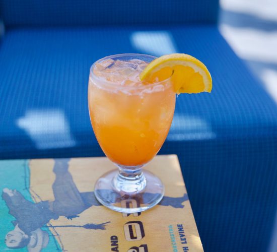 A refreshing orange drink garnished with a slice of orange sits on a table with a vintage book cover. The background features a blue cushioned seat, suggesting a relaxed outdoor setting at your favorite bar and grill.