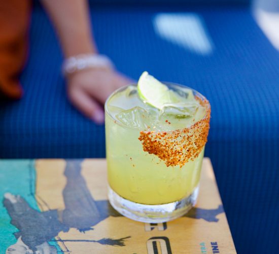 A close-up view of a cocktail in a glass with a salted and spiced rim, garnished with a lime wedge. The drink is on a colorful book cover featuring illustrations, with a person's arm resting on a blue, patterned cushion in the background—an experience you'd expect at the best restaurants and bars.