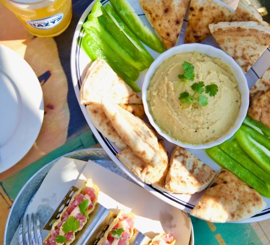 A platter of hummus surrounded by pita bread and green bell pepper slices, accompanied by a serving of taco-like appetizers filled with fresh ingredients, garnished with herbs. A glass of beer sits nearby on a colorful table often seen in trendy bars. Plates and utensils are partially visible.