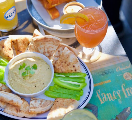 A close-up of a table at the best restaurant shows a variety of foods and drinks. There are pita slices and celery sticks around a bowl of hummus, a bowl of fried food sticks with dipping sauce, a glass of beer, a pink cocktail, and a green drink garnished with a lime wedge.