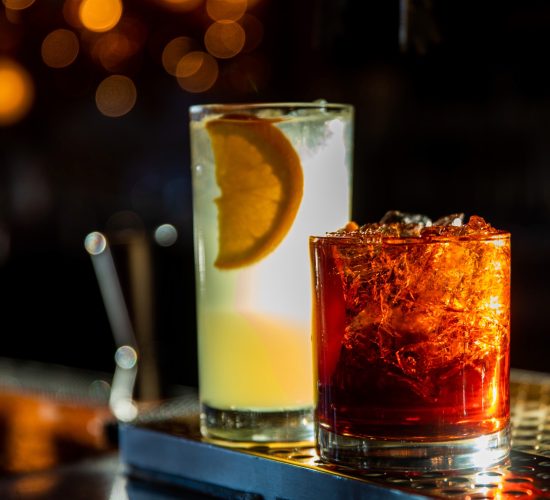 Two cocktail drinks on a bar counter at the popular bar and grill: a tall glass with a light-colored beverage garnished with an orange slice, and a shorter glass with a dark reddish-brown drink filled with ice. The background features soft, warm bokeh lights and promises of the best burger in town.