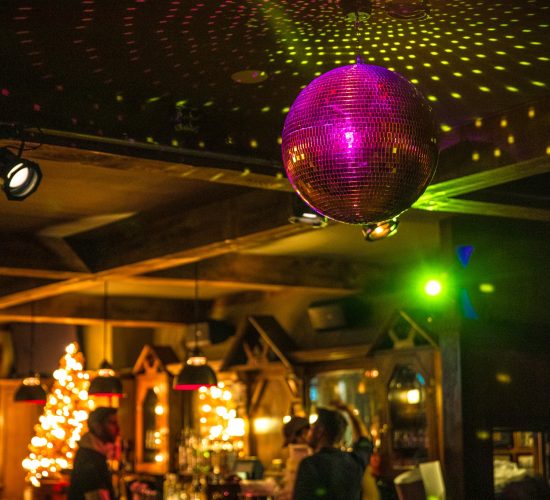 A warmly lit bar with Christmas trees decorated with lights in the background. A purple disco ball hangs from the ceiling, casting colorful light patterns around the room. The cozy bar and grill exudes a welcoming atmosphere, with people enjoying their cocktails and good company.