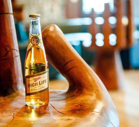 A bottle of Miller High Life beer rests on a large, sculpted wooden hand chair. The blurred background hints at cozy indoor seating in one of the best restaurants, with sunlight streaming through windows. The beer label displays the brand name and some text inviting craft cocktails nearby.