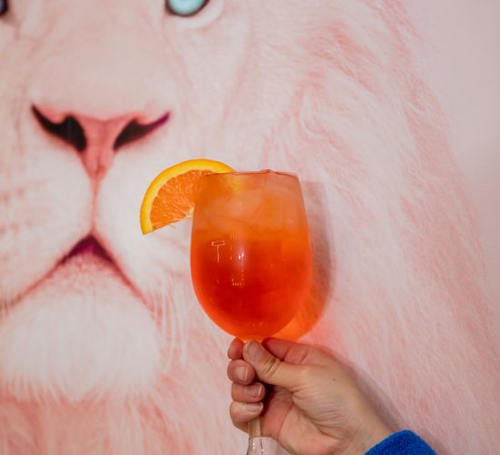 A hand in a blue sleeve holds a glass of iced orange cocktail, garnished with a slice of orange. The background features a close-up of a wall painting of a white lion with blue eyes, evoking the vibrant atmosphere often found in trendy bars and grills.