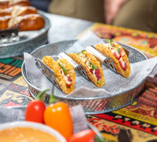 A metal plate holds four tacos, each in a taco holder, topped with various colorful ingredients and garnished with green herbs. The plate is lined with printed paper and placed on a colorful tabletop. In the foreground, red and yellow bell peppers are visible at this best restaurant known for both tacos and the best burger in town.