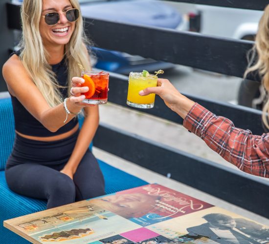 Two people are sitting at a table on an outdoor patio, clinking their cocktails in a toast. One is a woman with long blonde hair wearing sunglasses and smiling. The table is decorated with vibrant album covers, adding to the charm of the best restaurant and bar and grill in town. A blue chair and a wooden fence are visible.