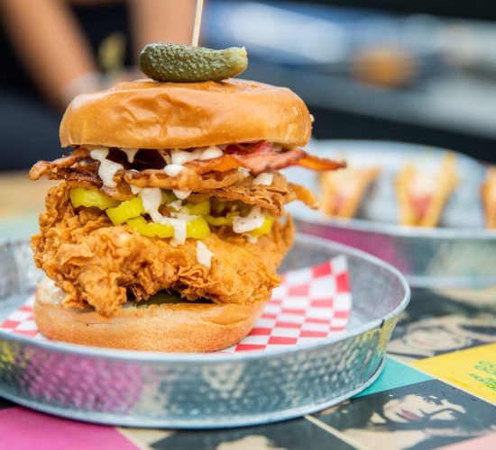 A crispy chicken sandwich topped with bacon, pickles, and a drizzle of sauce, skewered with a small pickle on top, served in a metal tray lined with red and white checkered paper. In the background of this bar and grill setting, there are additional small dishes and cocktails featuring lemon slices.