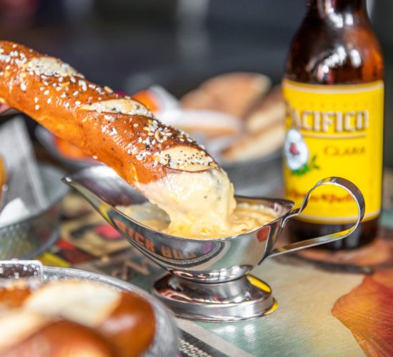 A pretzel partially dipped in a metal sauce cup filled with creamy cheese sauce sits on a tabletop, perfectly paired with a crisp Pacifico beer bottle positioned behind it. This mouth-watering scene, set against the blurred backdrop of a lively dining setting, hints at the best burger and cocktails offered by nearby bars.