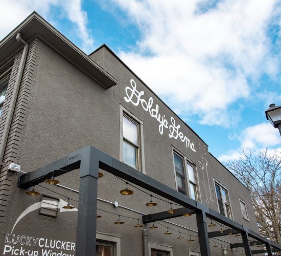 A building with a gray exterior under a blue sky with scattered clouds. The sign "Goldie’s Doma" is written in cursive on the upper part of the wall. Below it, there's a "Lucky Clucker Pick-up Window" sign. Known as the best restaurant, it’s perfect for enjoying cocktails under the nearby streetlamp's glow.