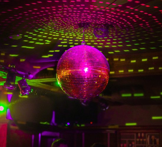 A vibrant disco ball hangs from the ceiling, casting colorful light reflections on the surrounding walls and ceiling. The room is dimly lit with a lively, festive atmosphere, suggesting a nightclub or party setting with patrons enjoying cocktails along with the beat.