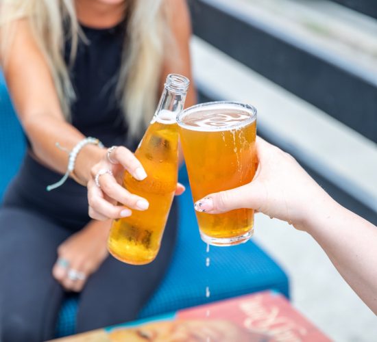 Two people toast with beer on a sunny day at the bar and grill. One holds a bottle while the other holds a glass. The person with the bottle has long blonde hair, sunglasses, and a black outfit, while the other person's hand is visible. They're seated at a table with a colorful tabletop.