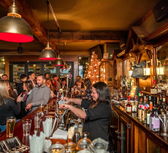 A lively bar scene with patrons engaged in conversation and cocktails. A bartender is seen mixing a drink behind the wooden bar. The bar and grill is decorated with a Christmas tree and festive lights. A variety of bottles and glasses are displayed, creating a warm, inviting atmosphere.