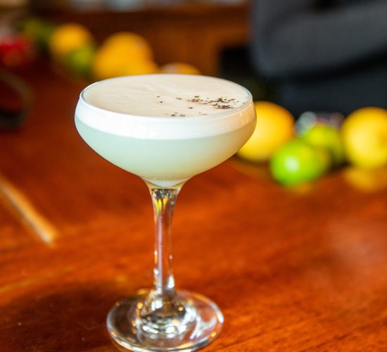 A cocktail glass filled with a creamy, white drink sits on a wooden table. The drink has a frothy top layer with a few dark specks of garnish. In the background, there are blurred fruits including limes and lemons, with a person partially visible, likely enjoying the best burger in town.