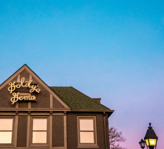 A two-story building with a sign reading "Goldy Gema" illuminated on its triangular roof. The building, known for having the best burger in town, features a green shingle roof and gray siding. A lamppost is visible on the right side, and the sky is clear with a gradient from blue to pink at sunset.