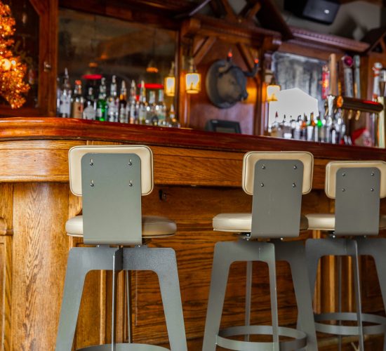 A wooden bar counter in a cozy pub and grill with five gray metal barstools. The bar is adorned with various bottles, glasses, and a few decorations, including a small, lit tree. Warm lighting illuminates the scene at the best restaurant in town, known for its welcoming atmosphere and best burgers.