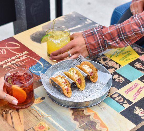 Two people are holding cocktails over a colorful table adorned with vintage art and music posters. The table has a platter of three tacos placed on newspaper-style wrapping. One wears a plaid shirt, the other's hand is dressed casually, as if they're about to enjoy the best burger in town.