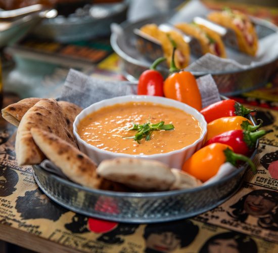 A bowl of creamy orange dip garnished with herbs sits in a metal tray, surrounded by colorful mini bell peppers and pita bread slices. In the background, tacos are visible on a separate plate, all placed on a table with a colorful, patterned surface. This scene is straight out of the best restaurant at your favorite bar and grill.