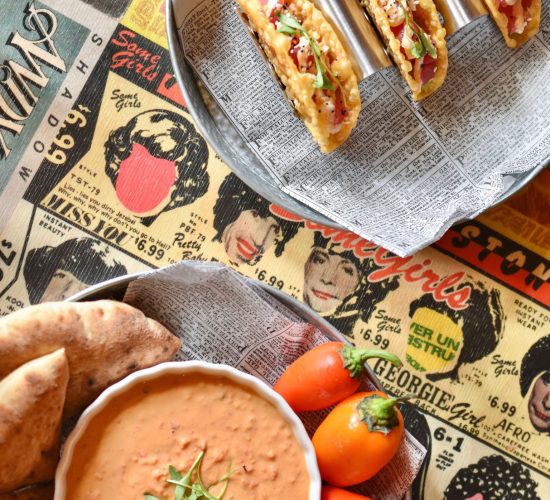 A plate of three tacos filled with various toppings, garnished with fresh herbs, sits above a round dish of dipping sauce surrounded by colorful bell peppers and sliced pita bread. The background features a vintage-style newspaper and comic design, perfect for one of the best restaurants in town.