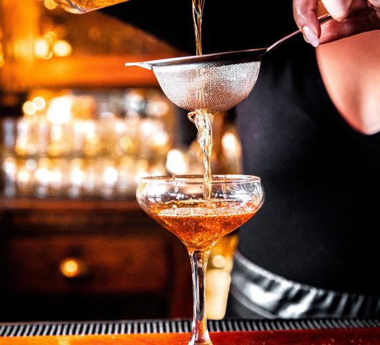 A bartender pours a golden-brown cocktail through a fine strainer into a stemmed glass. The warmly lit background showcases an array of bottles and the inviting wooden counter, reflecting the ambiance you’d expect from the best bars in town. The bartender’s hands and upper torso are visible.