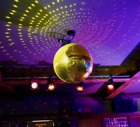 A golden disco ball hangs from the ceiling, illuminated by colorful spotlights. The lights create a pattern of reflections on the ceiling and walls, evoking a lively and festive atmosphere reminiscent of the best restaurant. Wooden beams and shelves are visible in the background, hinting at a cozy bar and grill.