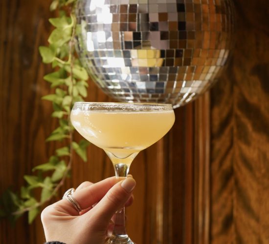 A hand with a dark sweater holds a coupe glass filled with a light yellow cocktail. A shiny disco ball hangs in the background, reflecting light, with a green leafy plant trailing beside it. The setting appears to be indoors at a cozy bar and grill, complete with a wooden backdrop.