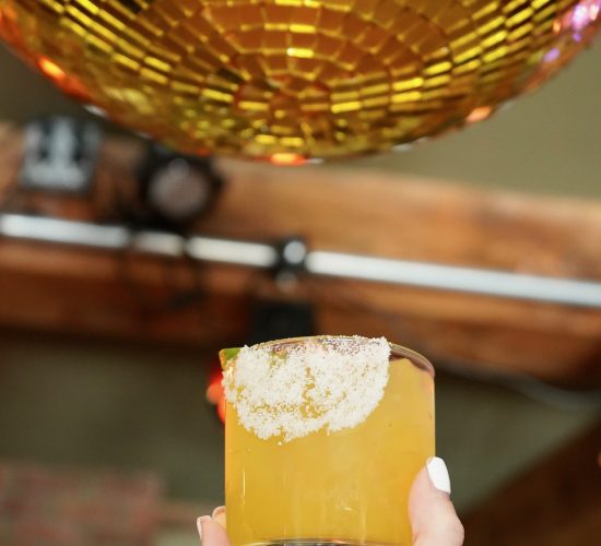 A hand with white nail polish holds up a glass of yellow cocktail with a sugared rim. The background features a large golden disco ball reflecting light. The setting appears to be a trendy bar or club with wooden beams and subtle lighting, perfect for enjoying some of the best cocktails in town.