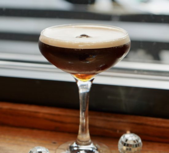 A glass filled with a dark, frothy cocktail sits on a wooden ledge by a window in a cozy bar and grill. Four small, silver disco ball decorations are scattered around the base of the glass, reflecting light. The background is slightly blurred, focusing attention on the drink and the disco balls.