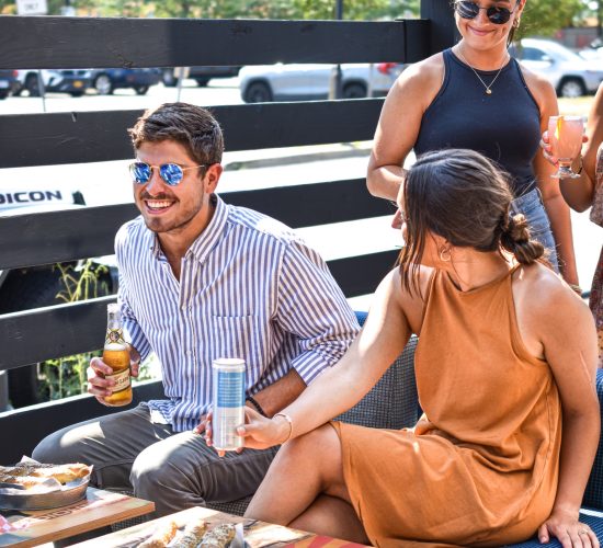 Three people are enjoying food and drinks outdoors on a sunny day. One man in a striped shirt and sunglasses holds a drink, while two women, one in a brown dress and the other in a black dress, are next to him. Snacks, drinks, and the best burger from the best restaurant are on the table in front of them.