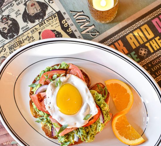 A hearty breakfast sandwich topped with a sunny-side-up egg, lettuce, and tomato is presented on a white plate. The plate also holds two slices of orange. The table, at one of the best restaurants in town, has a colorful collage of vintage magazine and poster art and a small lit candle.