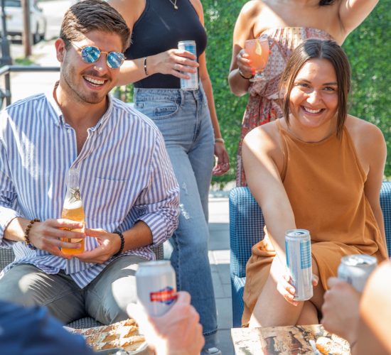 A group of five friends socializes outdoors at the best restaurant in town. Three women and two men enjoy drinks and snacks while sitting and standing around a small table. Everyone is smiling and appears to be having a good time in the sunny weather, with greenery in the background.