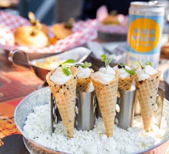 A silver tray holds ice cream cones filled with a savory dish, garnished with greens, and supported by a cone holder. The tray rests on a colorful tablecloth at this lively bar and grill, with blurred plates of burgers and a can of High Noon seltzer in the background.