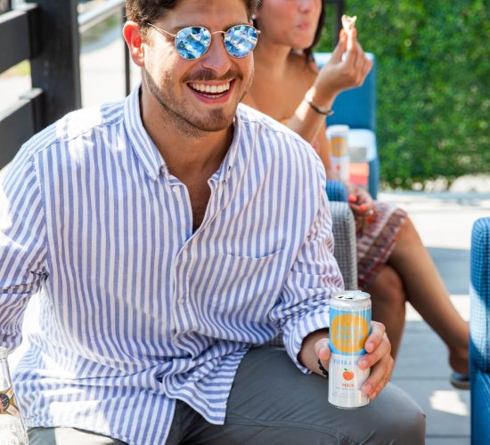 A man wearing sunglasses and a striped shirt enjoys a drink at an outdoor bar and grill. He is smiling and holding a can. In the background, a woman sits, holding a piece of food, also enjoying the meal. The table is filled with various dishes and drinks from the best restaurant in town.