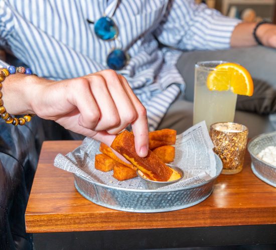 A person wearing a striped shirt and a beaded bracelet dips a fried cheese stick into sauce. Beside the dish, served on newspaper-print paper, sits a cocktail garnished with an orange slice and a candle holder, setting the perfect ambiance for enjoying bar snacks and sipping drinks.