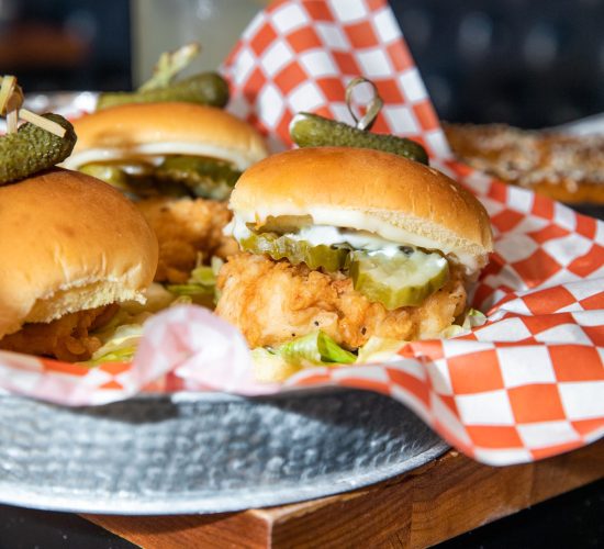 A close-up of sliders on a metal tray lined with red and white checkered paper. The sliders feature fried chicken topped with mayonnaise and pickle slices, with a pickle spear and toothpick securing each bun. In the blurred background, there’s a refreshing beverage hinting at the vibrant scene of a bar and grill.