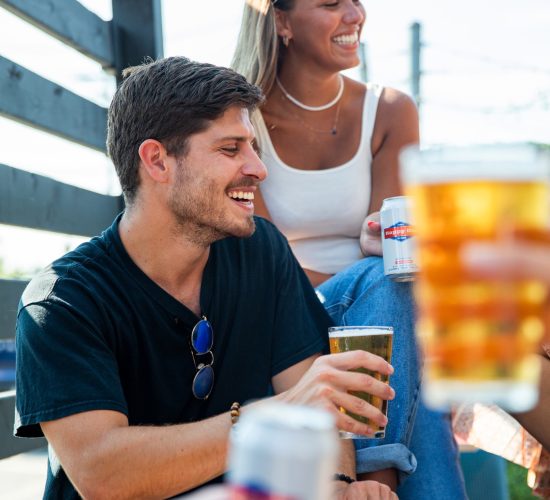 Two people, a man and a woman, are sitting outdoors at a bar and grill enjoying drinks. The man, in a black shirt and sunglasses, holds a beer, while the woman, in a white tank top, smiles warmly. They are engaged in conversation amidst the relaxed and cheerful atmosphere.